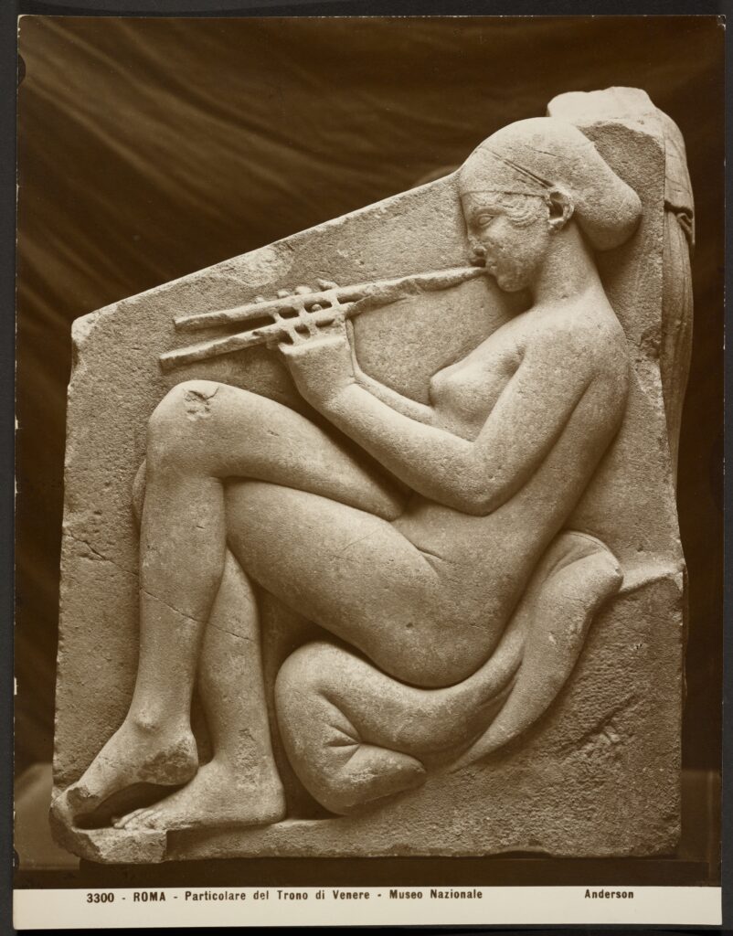 Photograph taken of a relief from the 'Throne of Venus' relief. A naked woman is depicted sitting while playing an aulos.