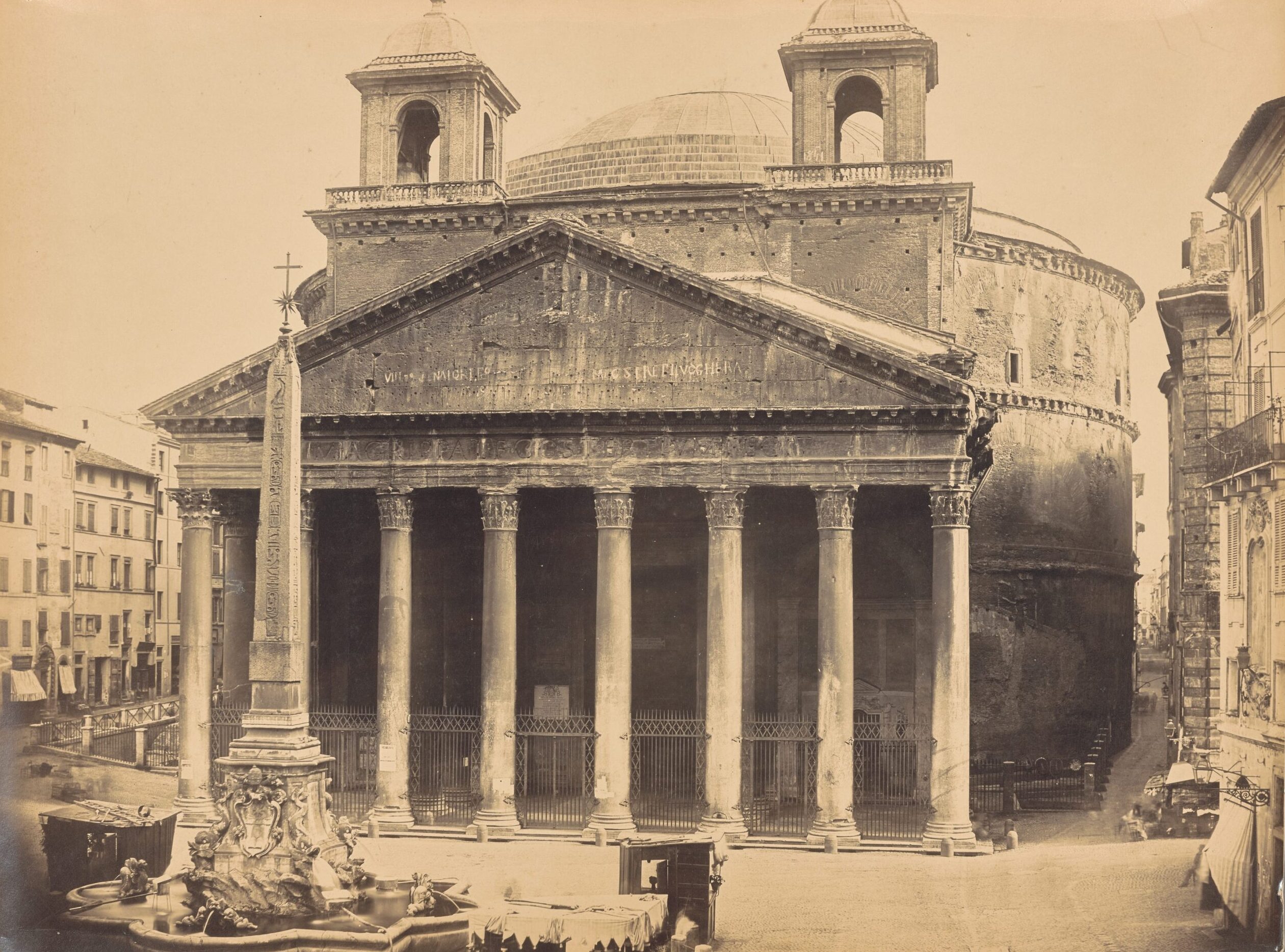 Exterior view of the Pantheon and the Fontana del Pantheon. The Pantheon is a circular building with a portico supported by eight Corinthian columns. There are two identical bell towers above the pediment. An Egyptian obelisk is in the center of the fountain in front of the Pantheon. Three kiosks are set up around the base of the fountain.